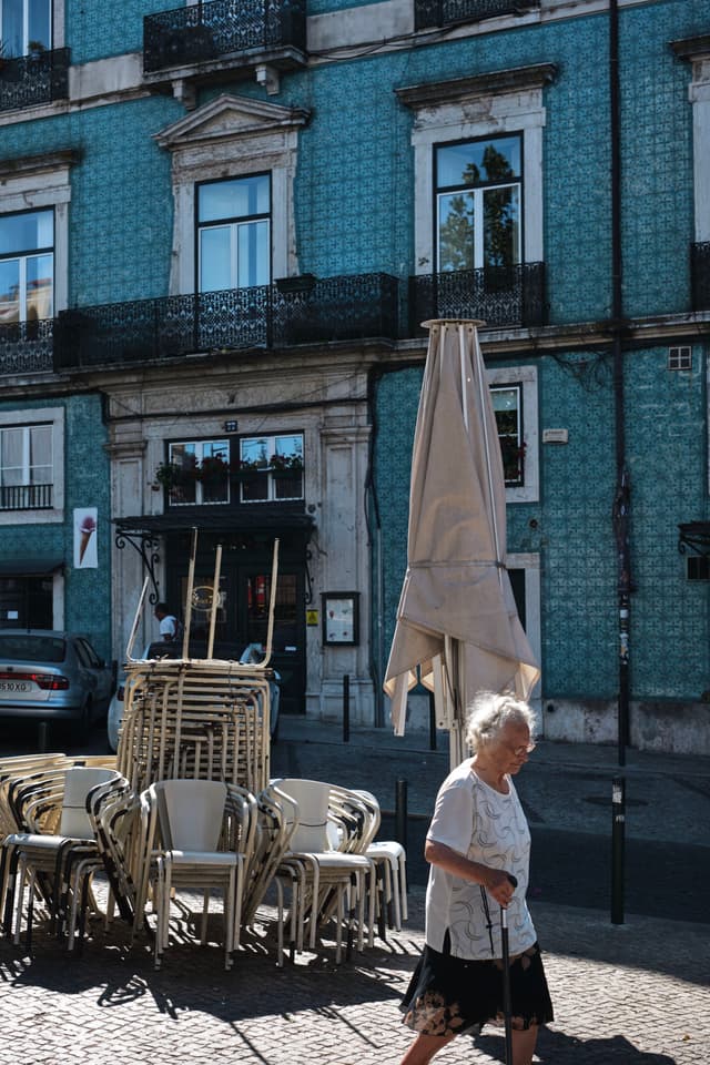 An elderly person walks past a stack of chairs and a closed umbrella on a cobblestone street, with a blue-tiled building in the background