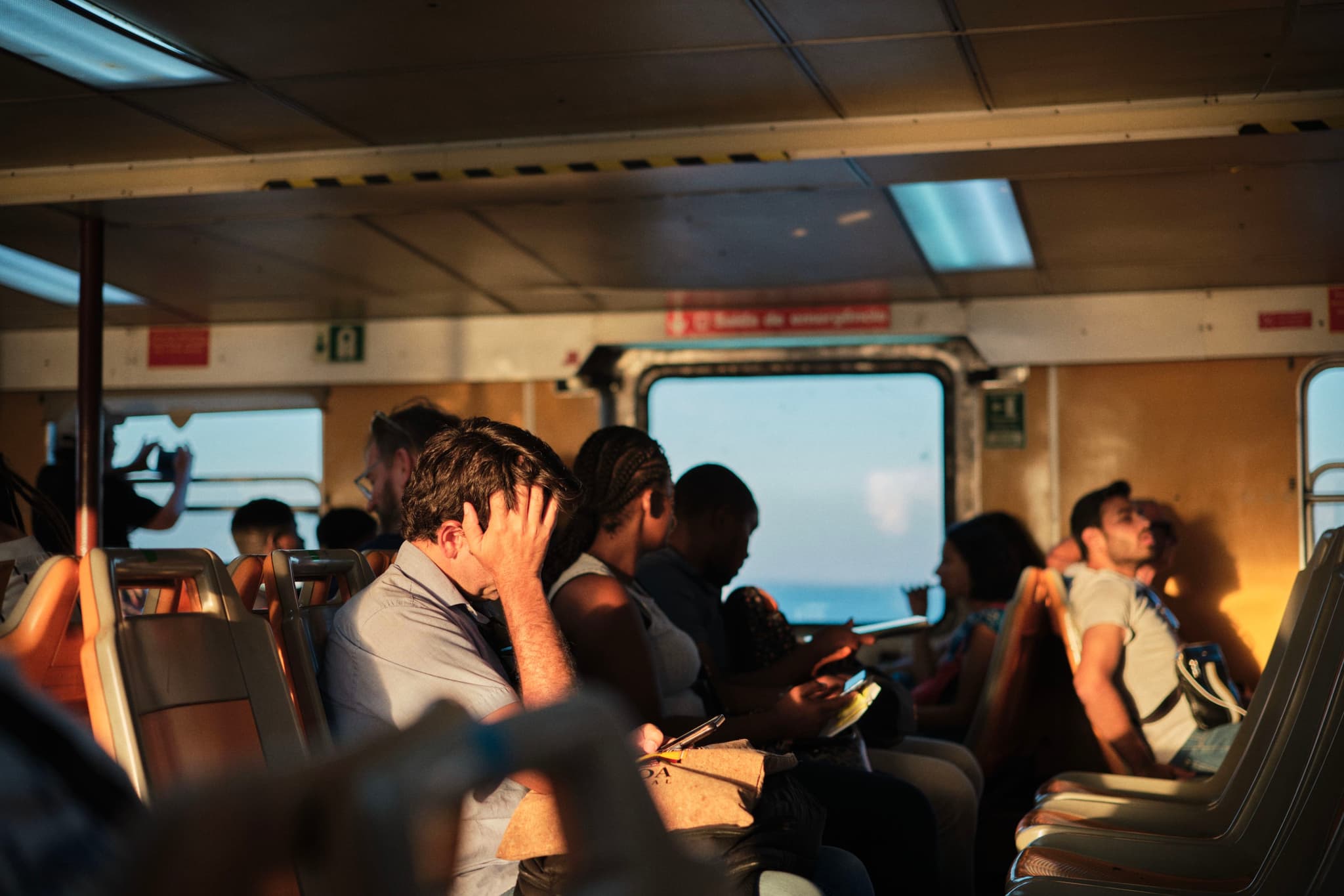 People sitting inside a ferry, some reading and others looking out the window, with sunlight streaming in
